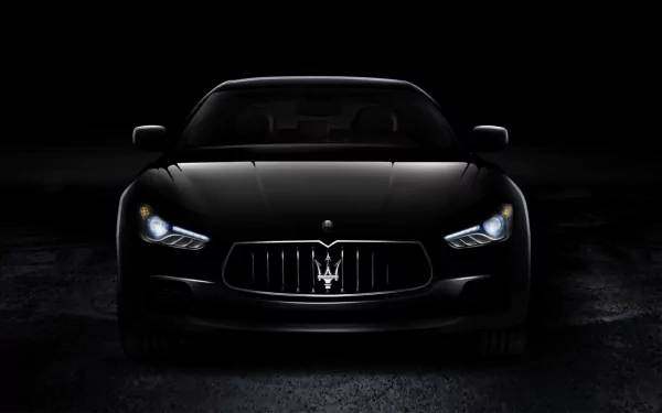 A sleek Maserati Ghibli in black, illuminated by its headlights, stands out against a dark background, showcasing the vehicle's elegant design in this HD desktop wallpaper.