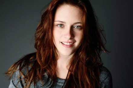 HD wallpaper featuring celebrity Kristen Stewart with tousled hair, wearing a gray top and smiling against a dark background.