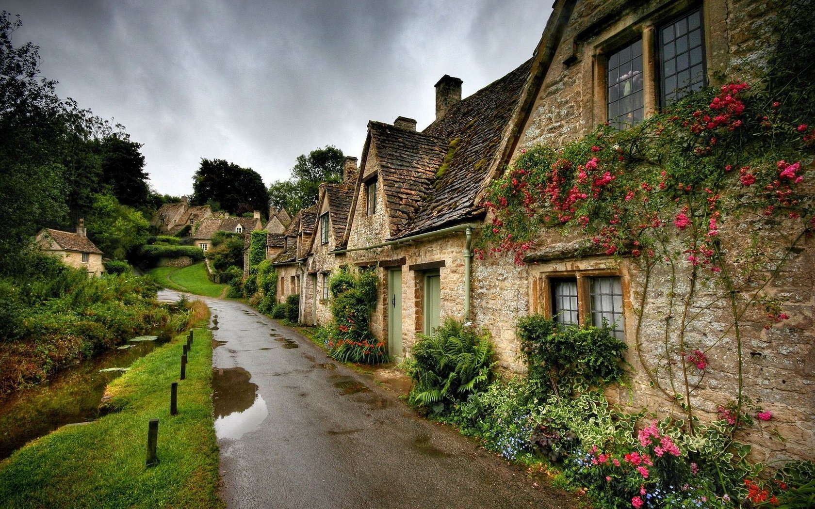 HD desktop wallpaper showcasing a man-made village scene with stone cottages, a winding wet road, and vibrant flowers under a cloudy sky.