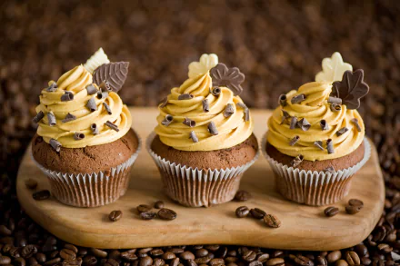 Delicious cupcakes with creamy frosting and chocolate decorations sit on a wooden platter, surrounded by coffee beans, creating a vibrant HD desktop wallpaper.