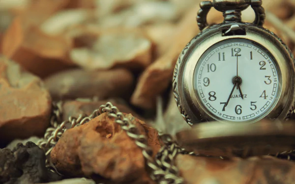 Close-up HD desktop wallpaper of a man-made antique pocket watch resting on scattered rocks with a detailed chain in a warm, earthy setting.