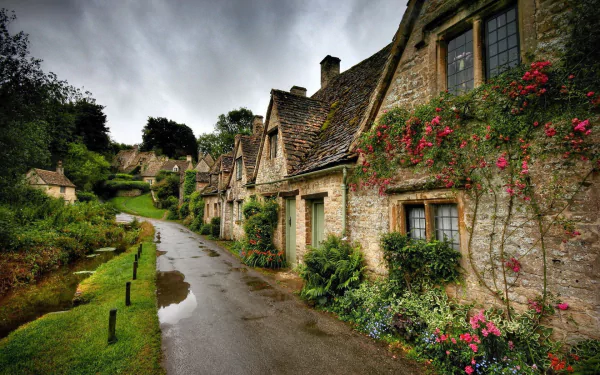 HD desktop wallpaper showcasing a man-made village scene with stone cottages, a winding wet road, and vibrant flowers under a cloudy sky.