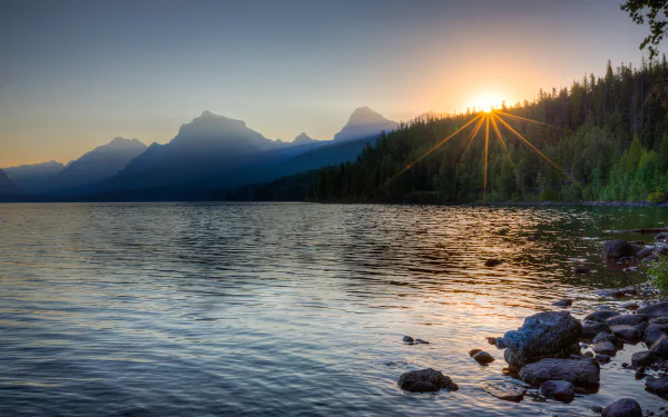 Sunset over Lake McDonald with calm waters, rocky shore, silhouetted trees, and clouds rolling above distant mountain peaks in this HD nature landscape wallpaper.