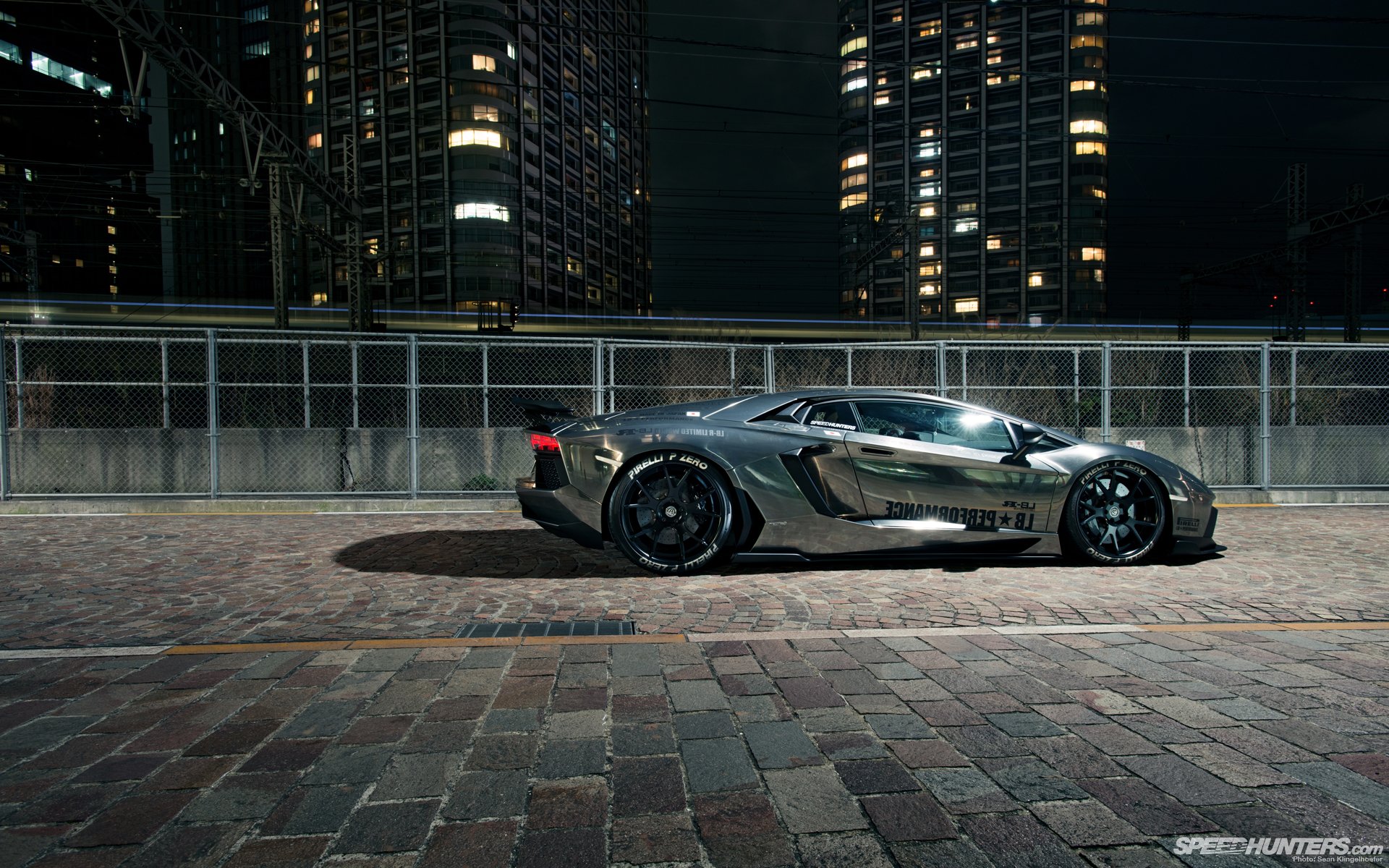 HD desktop wallpaper showcasing a sleek Lamborghini Aventador parked on a city street at night, with illuminated skyscrapers in the background.