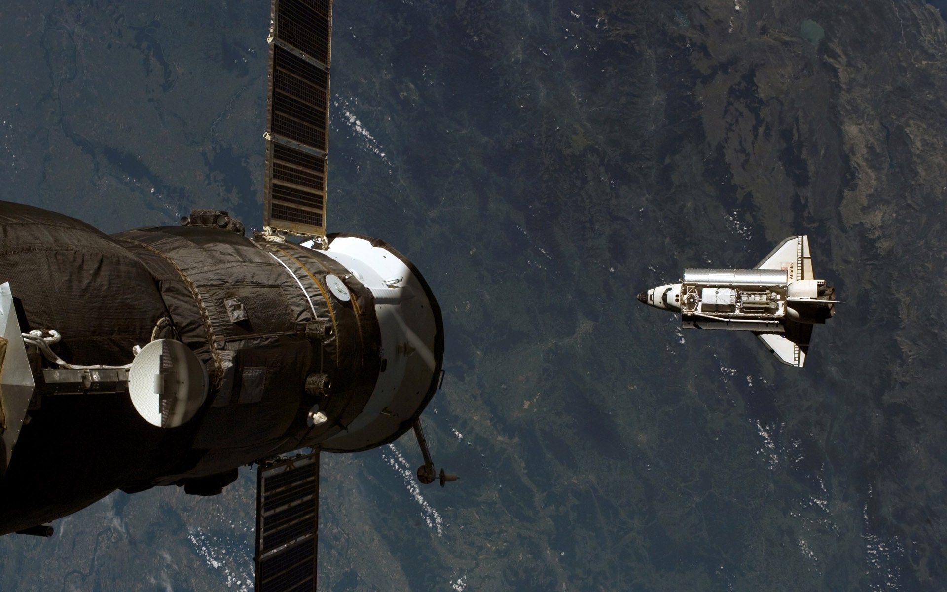 HD PC desktop wallpaper of vehicle Space Shuttle Atlantis approaching a spacecraft above Earth, with solar panels and the planet's surface visible beneath.