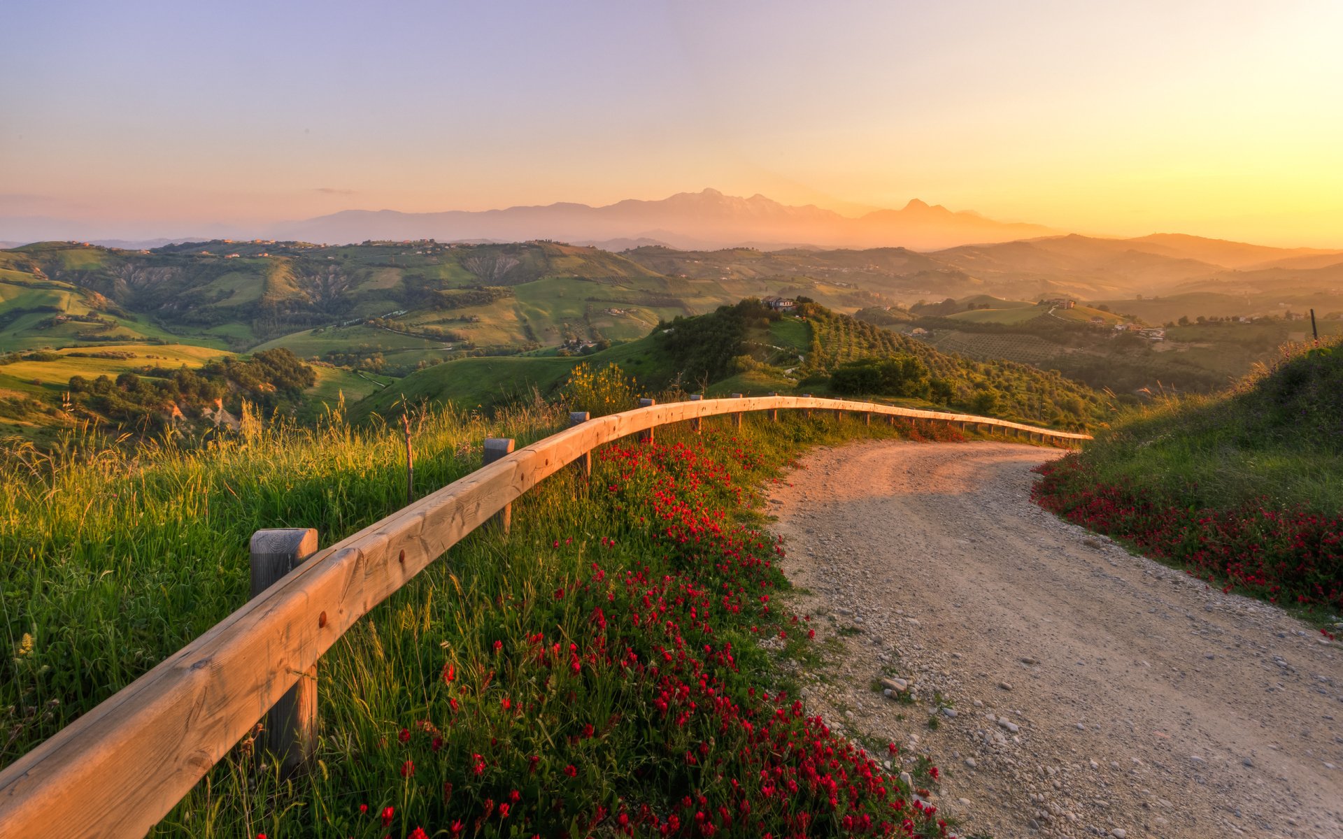 HD desktop wallpaper featuring a winding man-made road bordered by wooden railing and wildflowers, stretching through rolling hills at sunset.