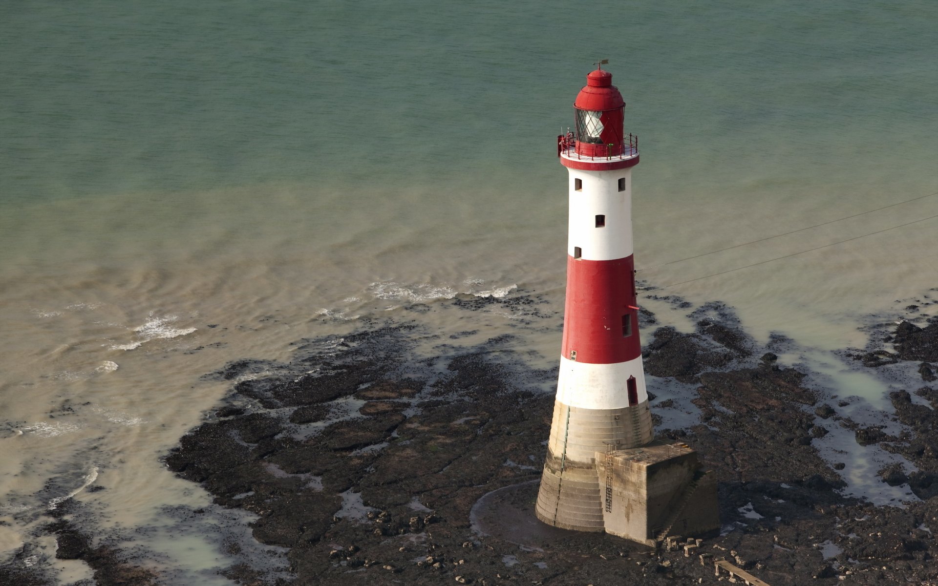 A striking red and white lighthouse stands on rocky shores, surrounded by calming waters, creating a captivating scene for a desktop wallpaper or background.