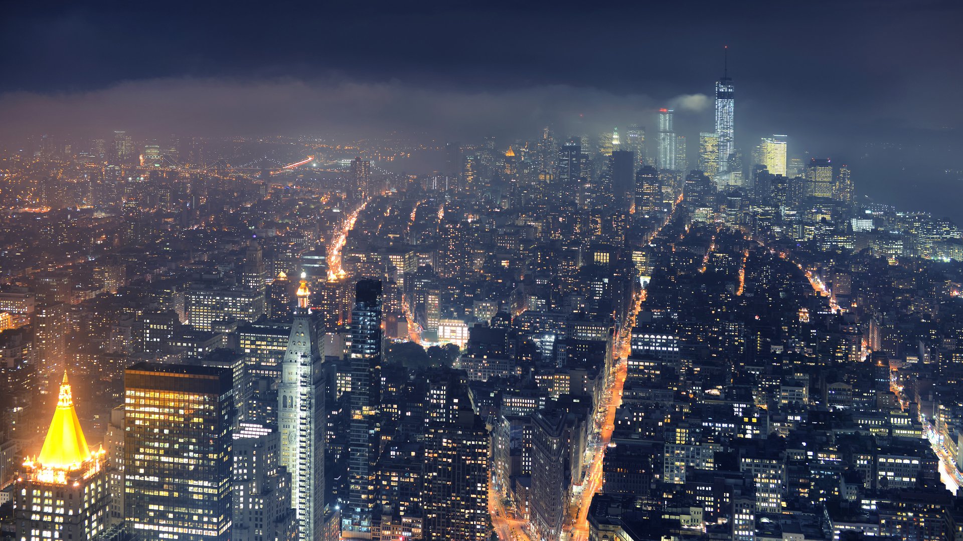 HD desktop wallpaper showing a night view of Manhattan, New York, with glowing city lights and skyscrapers under a cloudy sky.