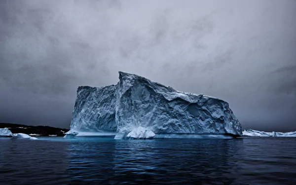 A striking iceberg looms majestically over dark waters beneath a moody sky, capturing the beauty of nature in this HD desktop wallpaper and background.