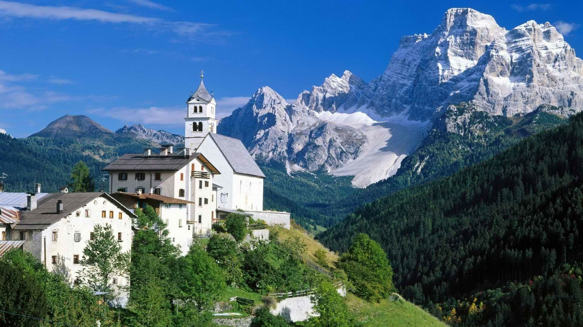 HD desktop wallpaper showing a picturesque man-made village nestled on a green hillside with towering snow-capped mountains in the background under a clear blue sky.