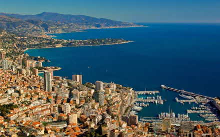 A stunning HD desktop wallpaper showing Monaco's cityscape and man-made harbor along the deep blue ocean, framed by distant mountains under a clear sky.