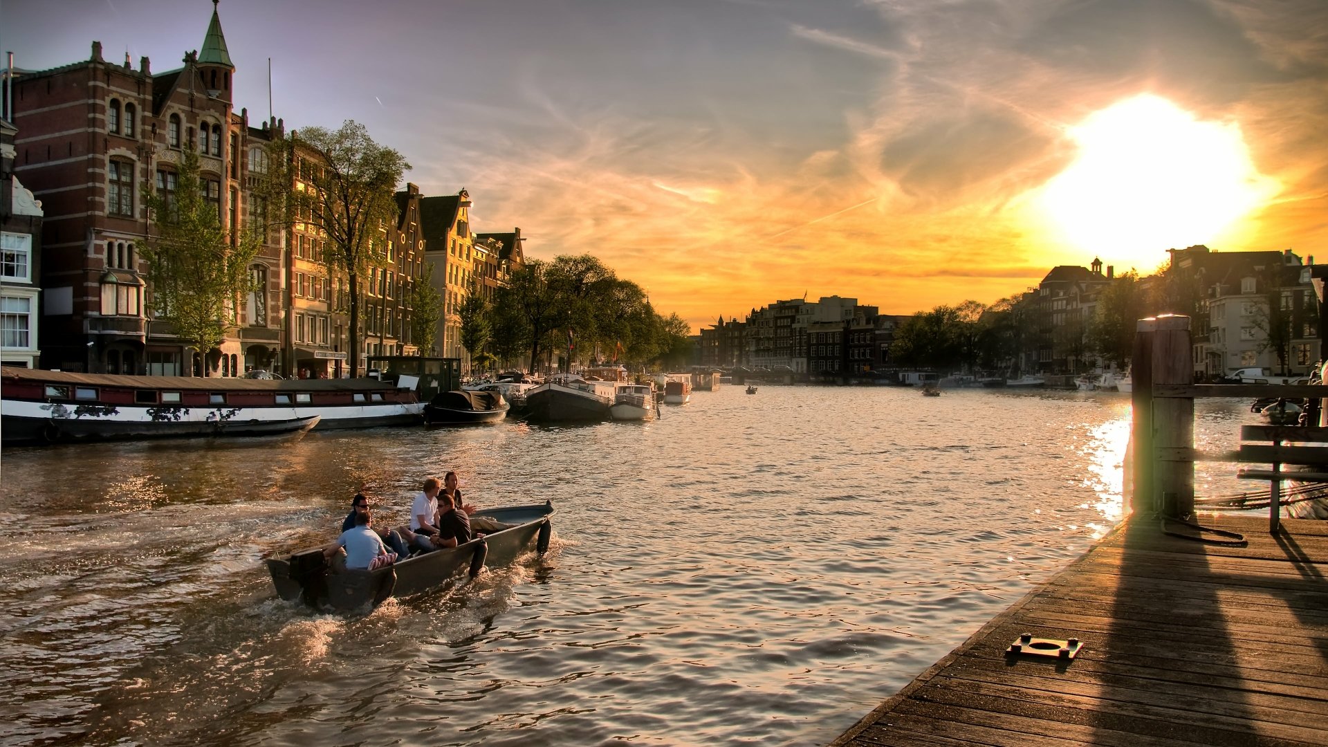 Sunset over Amsterdam's canal with historic buildings, boats on the water, and a wooden dock, captured in stunning 4K Ultra HD for a PC desktop background.