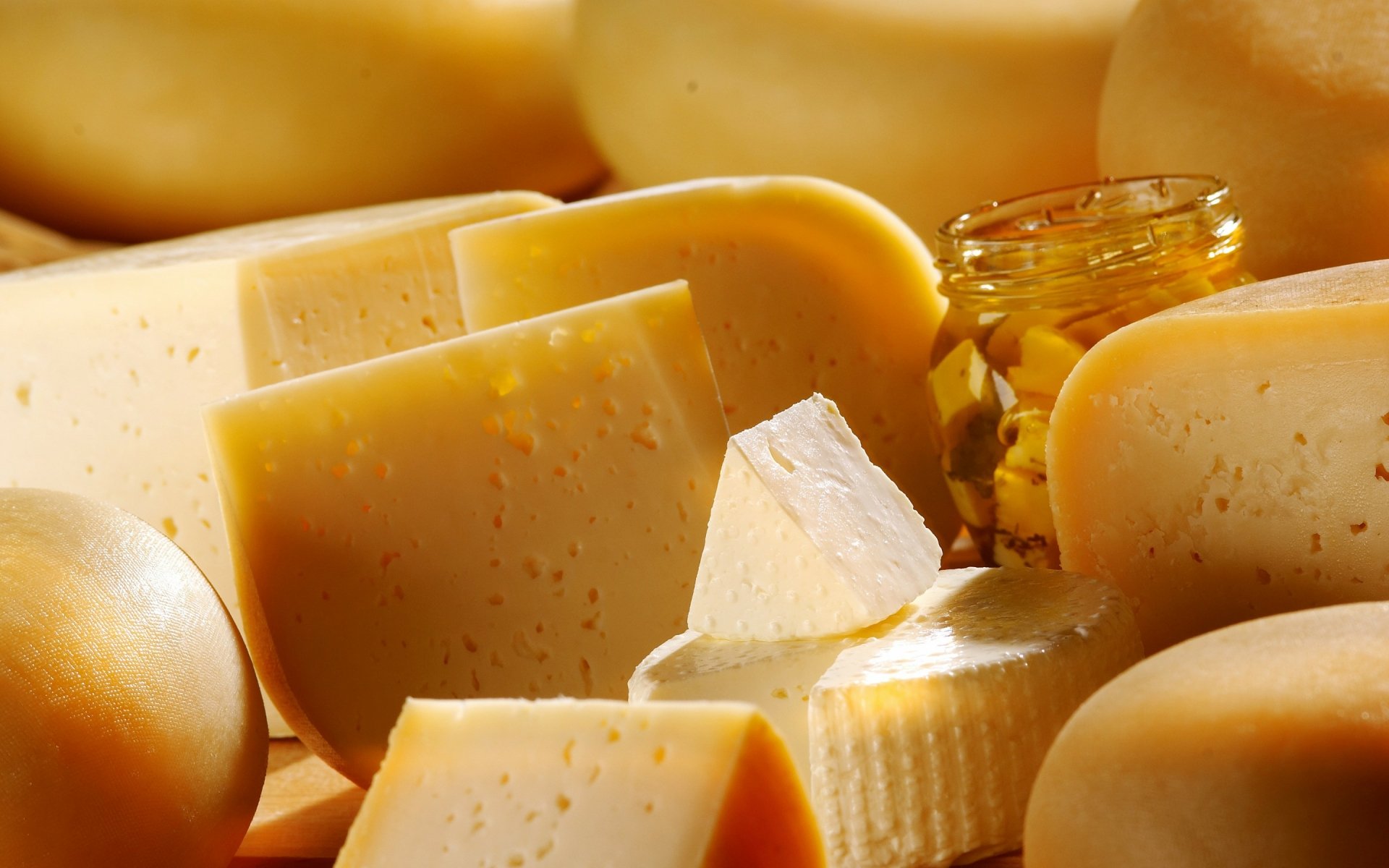 A vibrant assortment of cheese varieties, including blocks and wedges, accompanied by a jar of pickled items, set on a wooden surface. A stunning HD wallpaper for food enthusiasts.