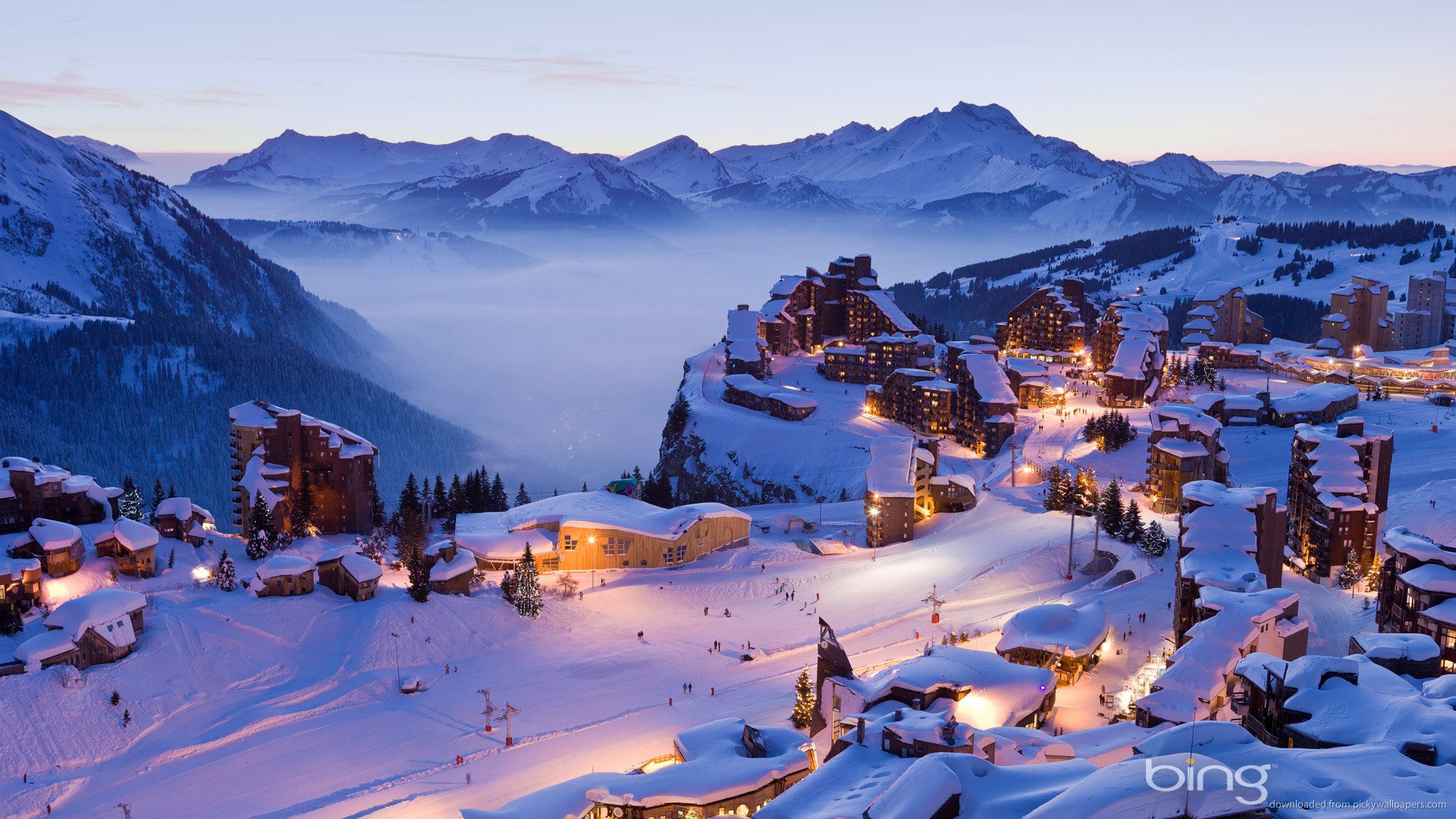HD desktop wallpaper featuring a snow-covered mountain village at dusk, with illuminated buildings nestled among rugged peaks under a clear sky.