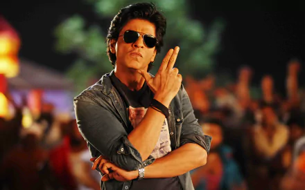 A stylish Bollywood actor poses confidently, wearing sunglasses and a casual outfit, set against a vibrant background full of energy. This HD wallpaper features Shah Rukh Khan.