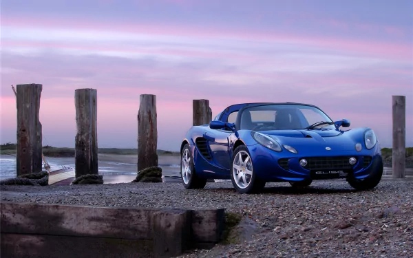 A sleek blue Lotus Elise sports car parked on a rocky shore at sunset, with wooden posts and calm water in the background, captured as an HD desktop wallpaper.