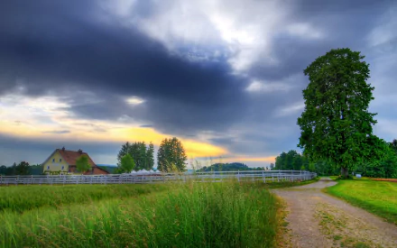 A serene landscape featuring a tall tree beside a winding path, with a farm and lush fields under a dramatic sky, capturing the beauty of nature in a man-made setting.