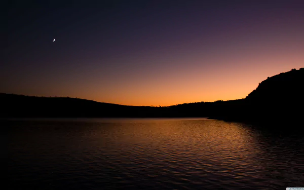 A serene lake at twilight, reflecting the vibrant hues of sunset, with a crescent moon visible in the sky. This nature scene is captured in stunning 4K Ultra HD quality.