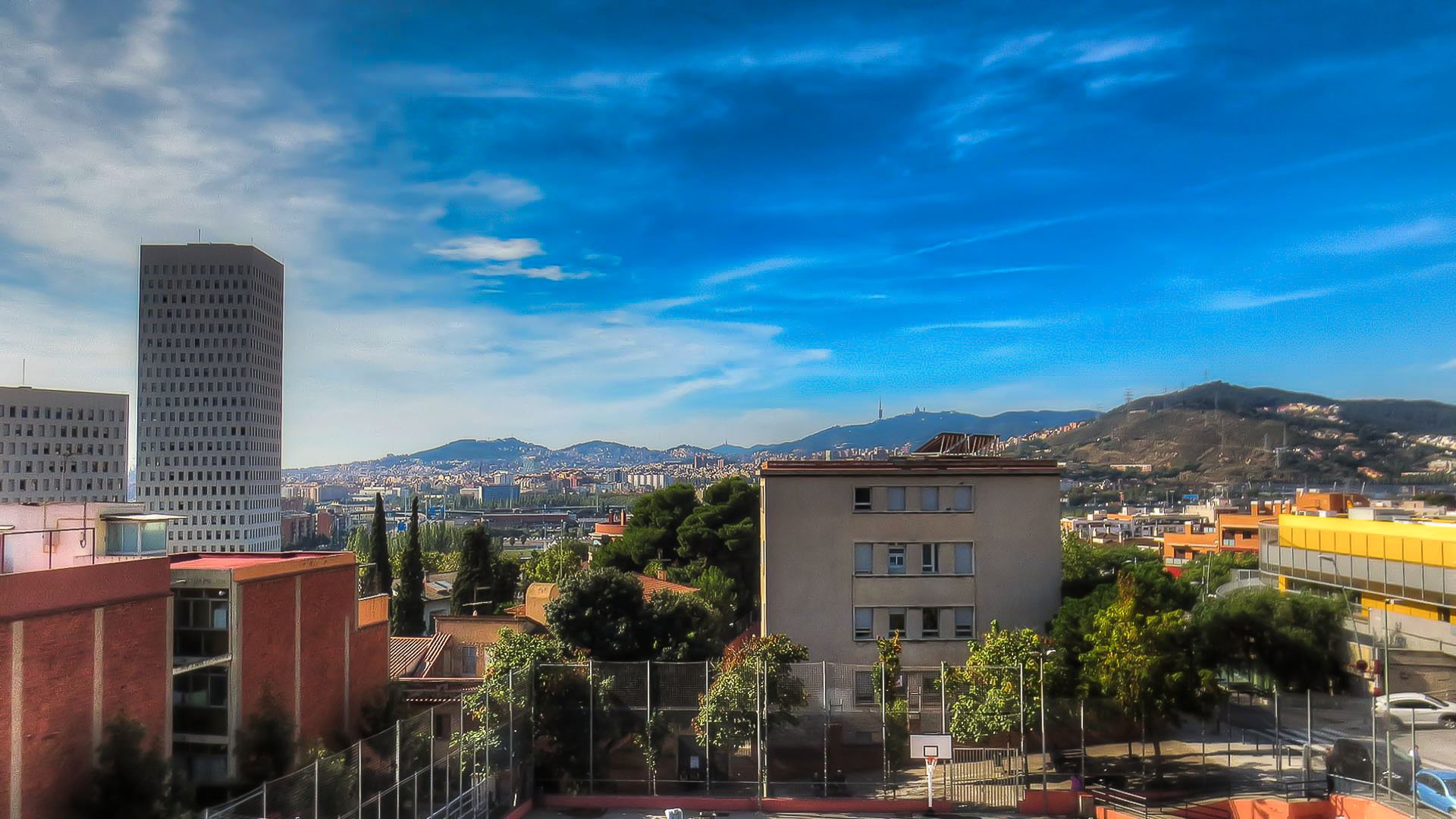 HD desktop wallpaper showcasing a man-made cityscape of Barcelona with buildings, greenery, and mountains under a vibrant blue sky.