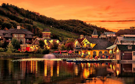 HD PC desktop wallpaper and background: a man-made lakeside village at sunset, illuminated cottages and waterfront restaurants reflecting in calm water below rolling green hills.