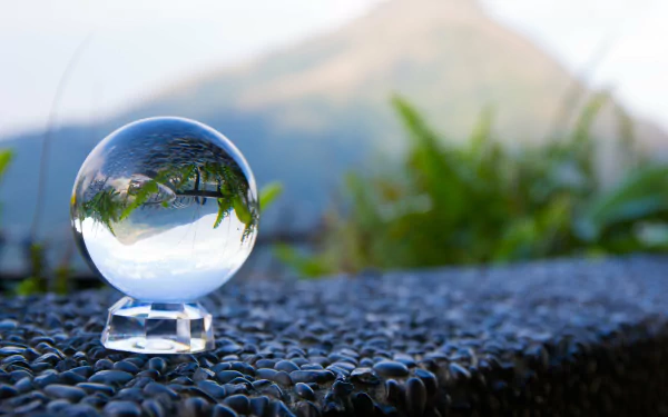 A glass globe sits on a textured surface, reflecting a serene landscape with greenery and mountains in the background. This high-definition image serves as an elegant desktop wallpaper.