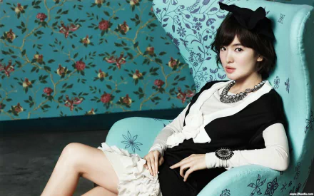 A stylish Korean actress sits elegantly in a vibrant blue chair, adorned with floral wallpaper, showcasing a blend of sophistication and charm in this HD desktop wallpaper.