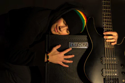 HD desktop wallpaper showing a person embracing a Fender bass guitar amplifier, with a black bass guitar beside them in a dimly lit music-themed scene.