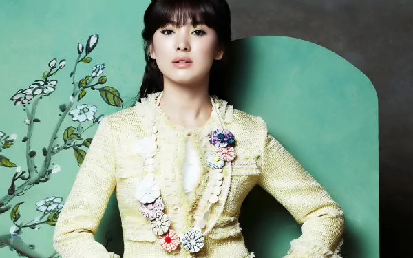 A stylish HD wallpaper featuring Korean actress Song Hye Kyo, showcasing her elegant outfit and poised expression against a vibrant green backdrop with floral accents.