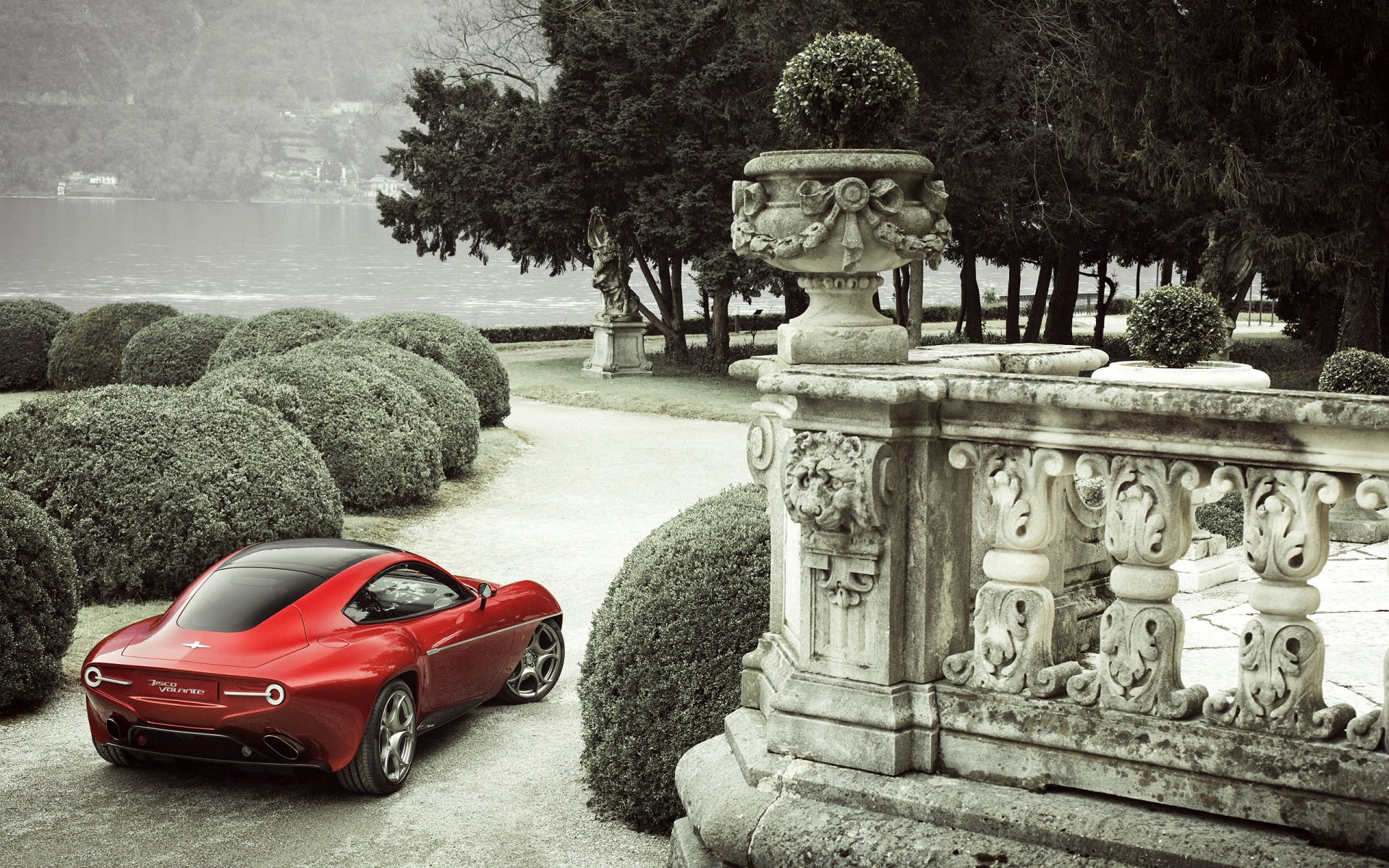 Red Alfa Romeo Disco Volante vehicle parked on an ornate lakeside terrace with topiary and stone balustrade — 2K Quad HD PC desktop wallpaper/background.