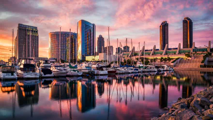 A stunning 4K Ultra HD view of San Diego's skyline at sunset, featuring man-made marina structures and boats reflecting on calm water.