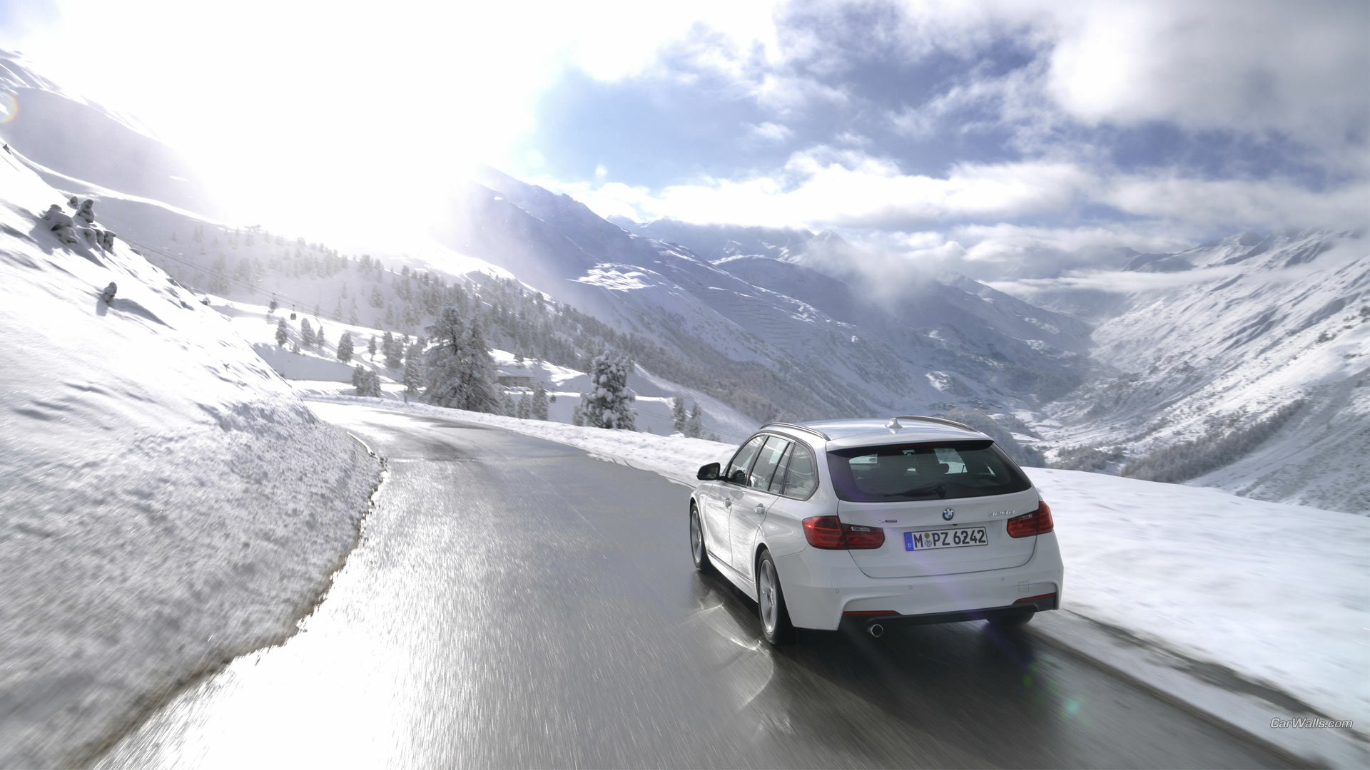 HD PC desktop wallpaper and background: a white 2013 BMW 320d driving on a wet mountain road through a snowy alpine landscape under a dramatic cloudy sky.