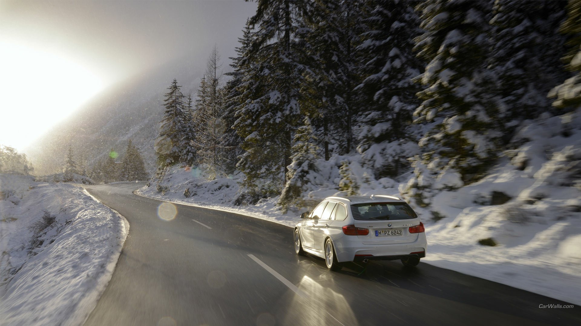 2013 BMW 320d driving along a wet, winding mountain road lined with snow-covered pines and bright sun glare — HD PC desktop wallpaper/background.