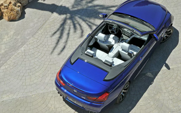 HD desktop wallpaper featuring a top-down view of a blue BMW M6 Convertible parked on a sunlit stone driveway with shadows of palm trees.