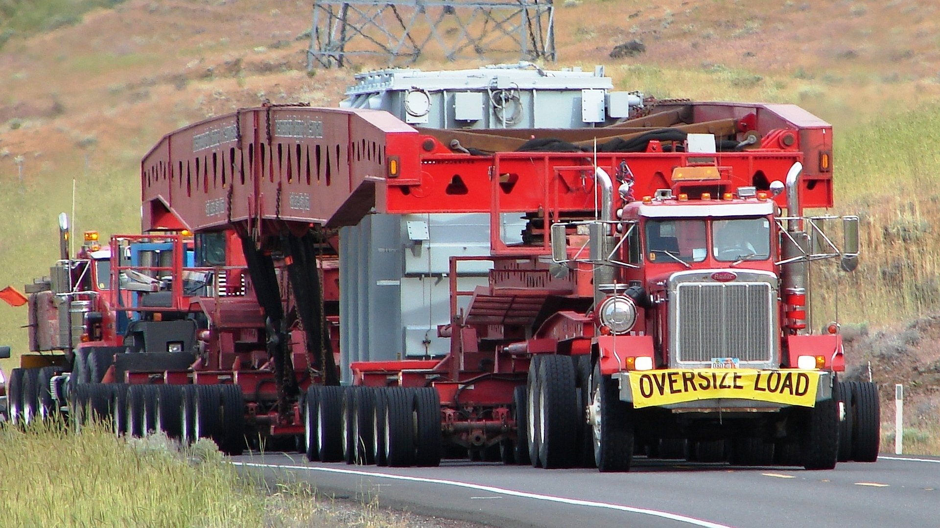 HD desktop wallpaper featuring a red Peterbilt vehicle transporting an oversized load on a highway through a rural landscape.