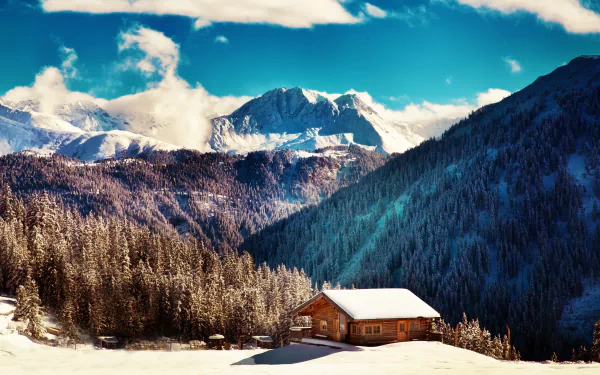 A serene cabin nestled in a snow-covered landscape, surrounded by majestic mountains and evergreen trees, captured in stunning 4K Ultra HD for a captivating desktop wallpaper.