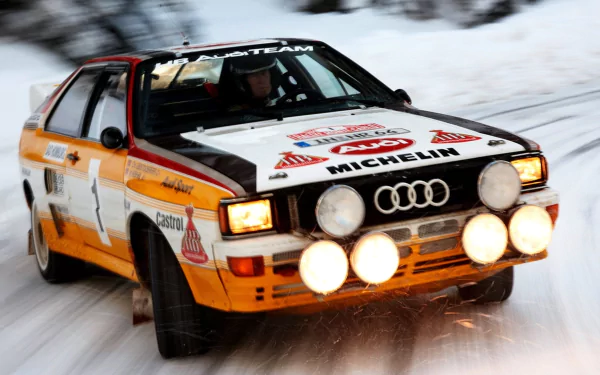 HD PC desktop wallpaper of an Audi Quattro rally car racing through snow, headlights and four auxiliary spotlights blazing.