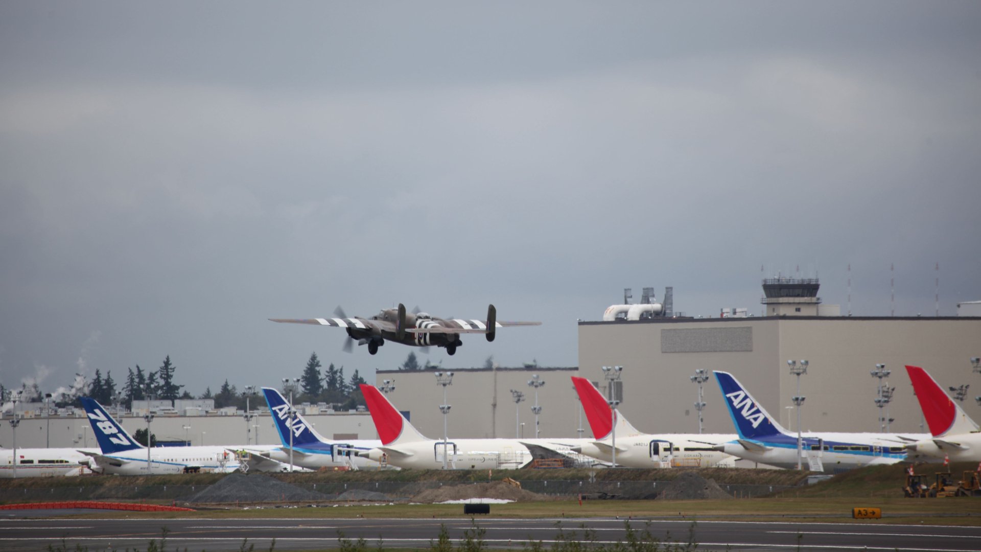 HD desktop wallpaper showing a North American B-25 Mitchell military aircraft flying low over an airport with commercial planes and a control tower in the background.