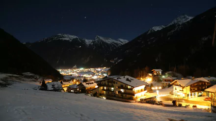 A serene night view of Mayrhofen, Austria, showcasing charming lit buildings against a backdrop of snow-covered mountains under a starry sky. 4K Ultra HD quality wallpaper.