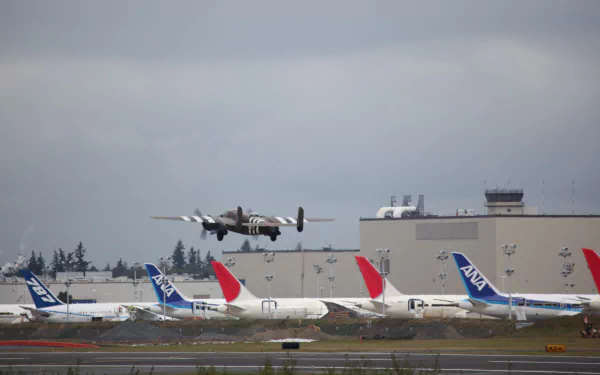 HD desktop wallpaper showing a North American B-25 Mitchell military aircraft flying low over an airport with commercial planes and a control tower in the background.