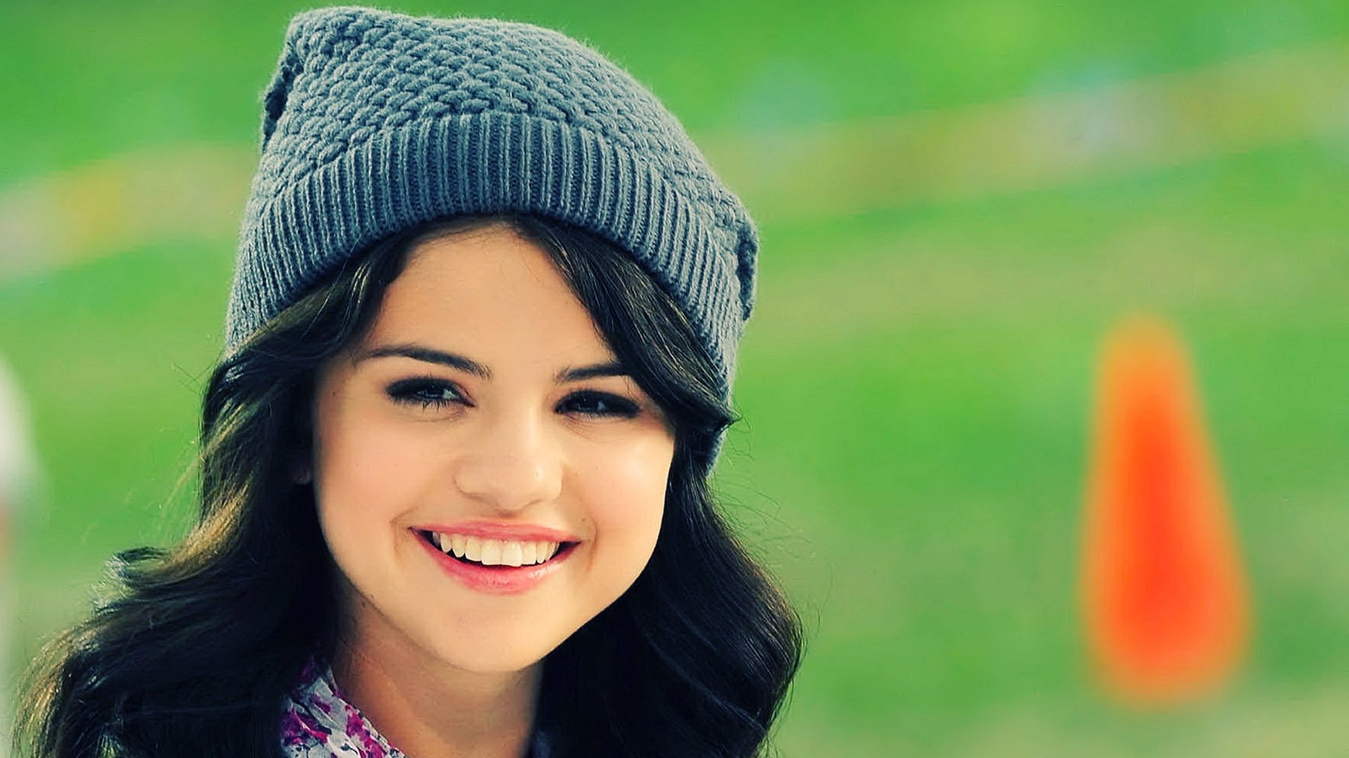 HD desktop wallpaper featuring Selena Gomez, smiling and wearing a gray beanie, set against a green background. A vibrant pop of color adds energy to the scene.