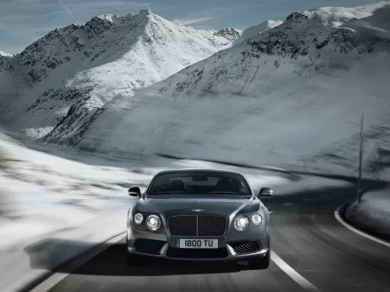HD desktop wallpaper featuring a Bentley Continental GT V8 driving on a winding mountain road with snow-covered peaks in the background.