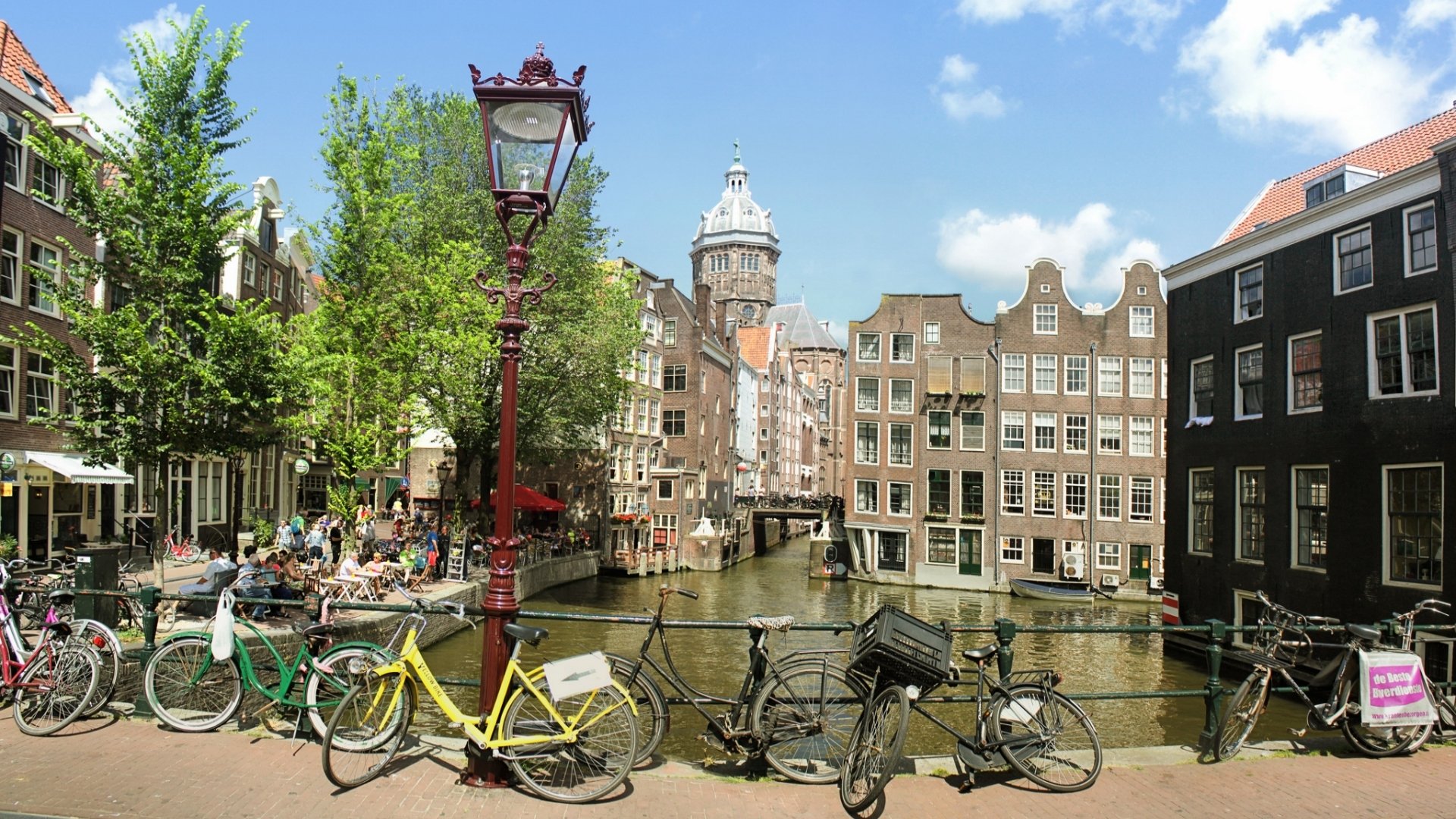 HD desktop wallpaper showcasing a man-made canal scene in Amsterdam with bicycles lined along the railing and traditional Dutch buildings under a bright blue sky.