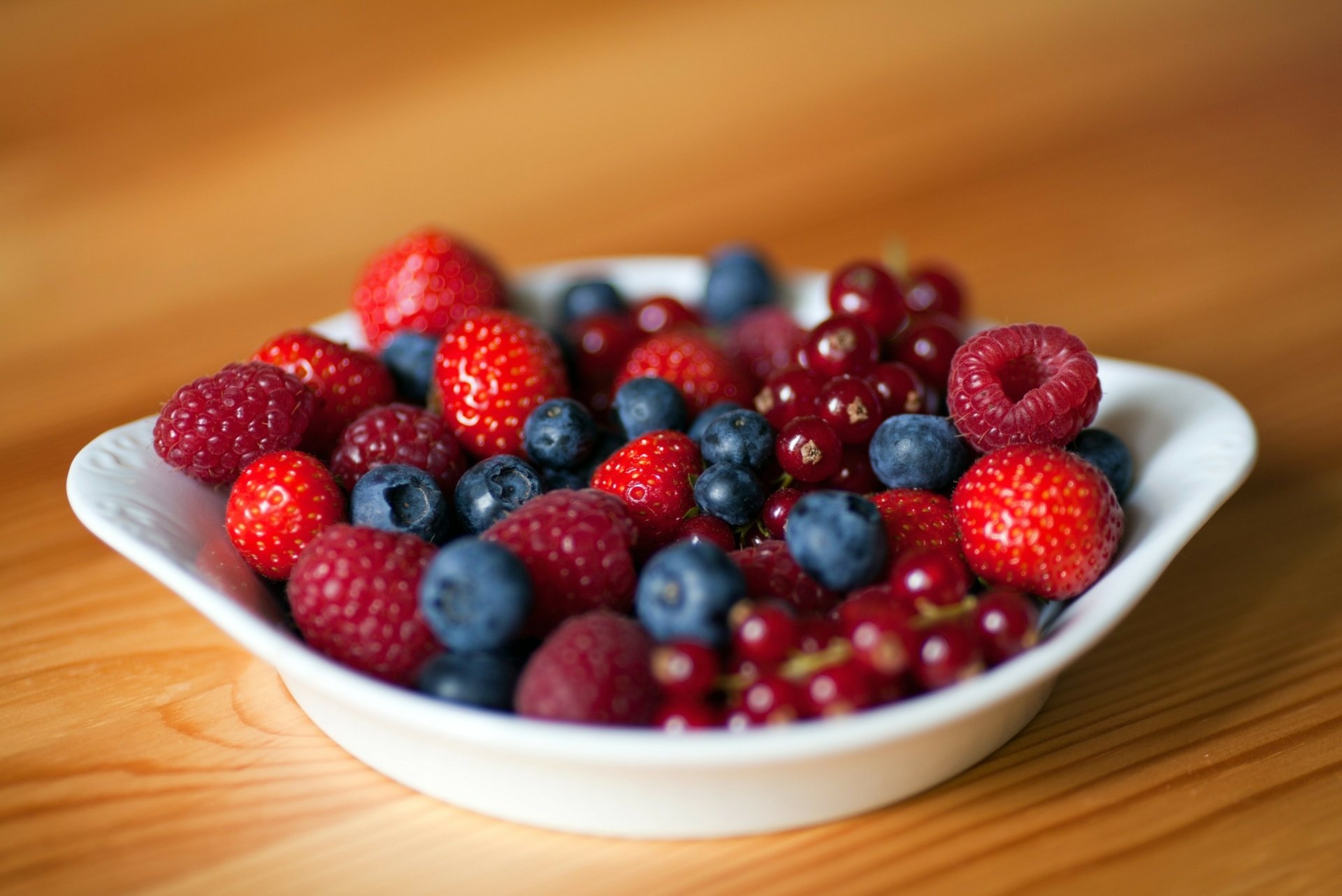 HD PC desktop wallpaper featuring a close-up of a white bowl filled with fresh strawberries, raspberries, blueberries, and red currants on a wooden surface.