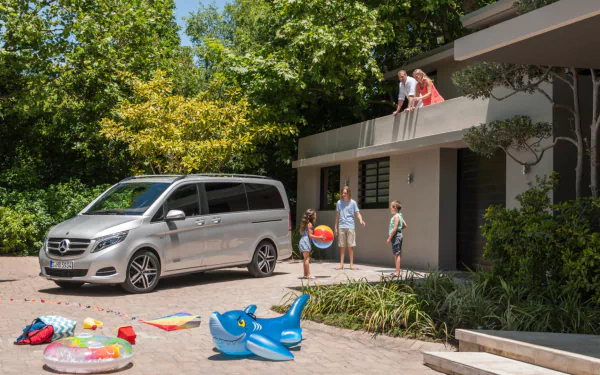 HD PC desktop wallpaper and background: silver Mercedes‑Benz V‑Class vehicle parked by a modern home while a family plays with beach balls and inflatables in the sunlit driveway.