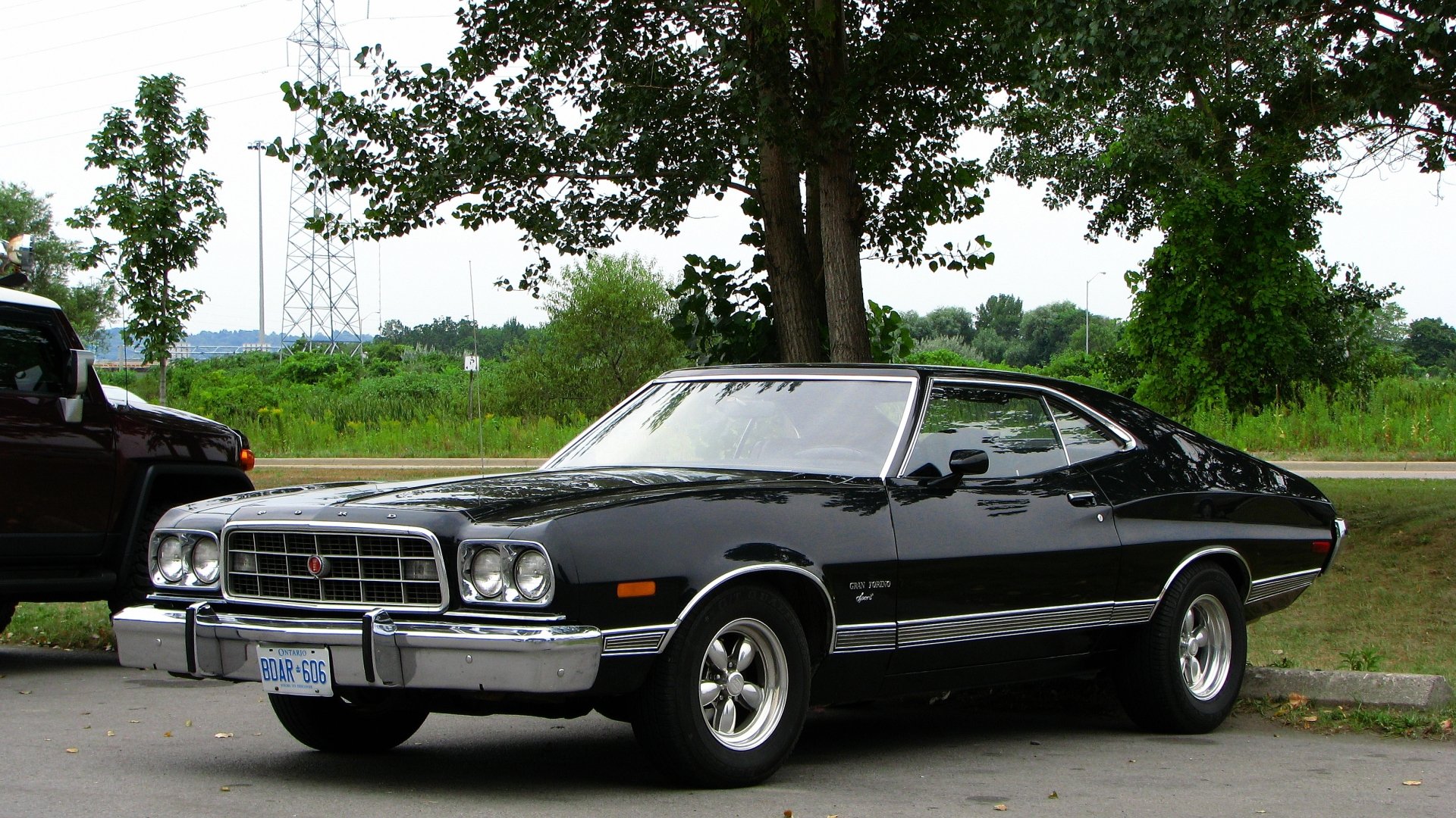 HD PC desktop wallpaper featuring a classic black Ford Gran Torino parked by a tree with greenery and a clear sky in the background.