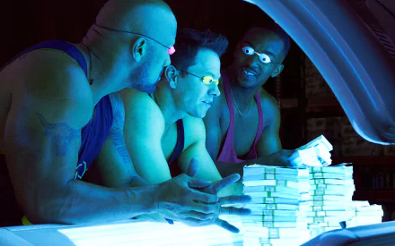Three muscle-bound men, Dwayne Johnson, Mark Wahlberg, and Anthony Mackie, inspect stacks of cash under glowing lights in a scene from the movie Pain & Gain.
