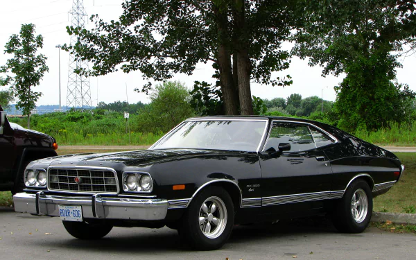 HD PC desktop wallpaper featuring a classic black Ford Gran Torino parked by a tree with greenery and a clear sky in the background.