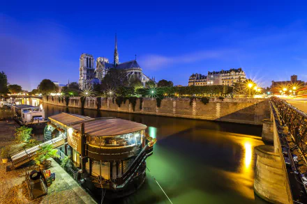 A stunning HD desktop wallpaper of Paris at night, featuring a riverside view with a boat and the iconic Notre-Dame Cathedral illuminated against a deep blue sky.