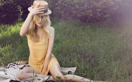 HD PC desktop wallpaper of a French actress/celebrity in a yellow sundress seated on a blanket in sunlit grass, tilting a straw hat over her face amid soft, dreamy lighting.