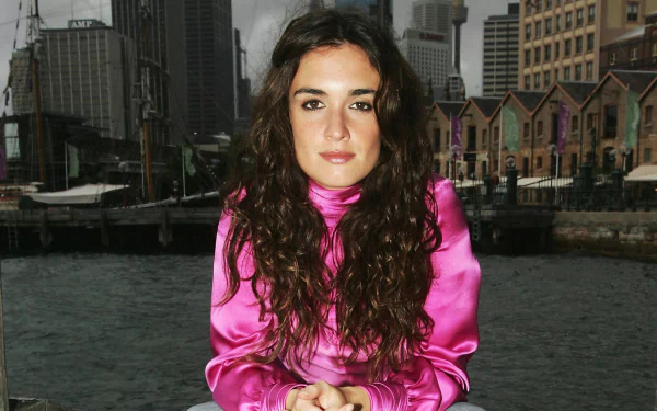 HD desktop wallpaper: Spanish actress and celebrity in a pink satin blouse, seated by a waterfront with a city skyline behind her.